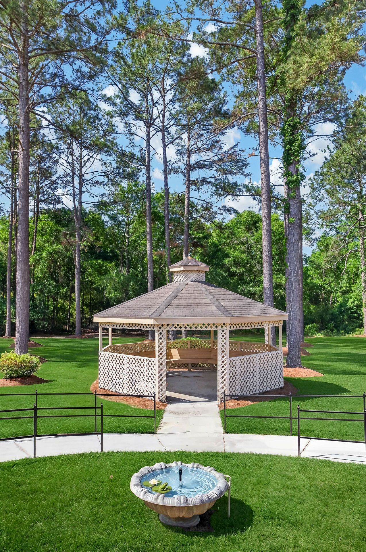 Gazebo in a landscaped park with green lawn, trees, and a small fountain in the foreground