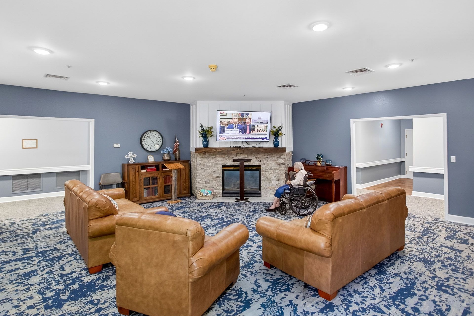 Cozy living room with tan chairs, blue patterned carpet, fireplace, and wall-mounted TV built above the mantel