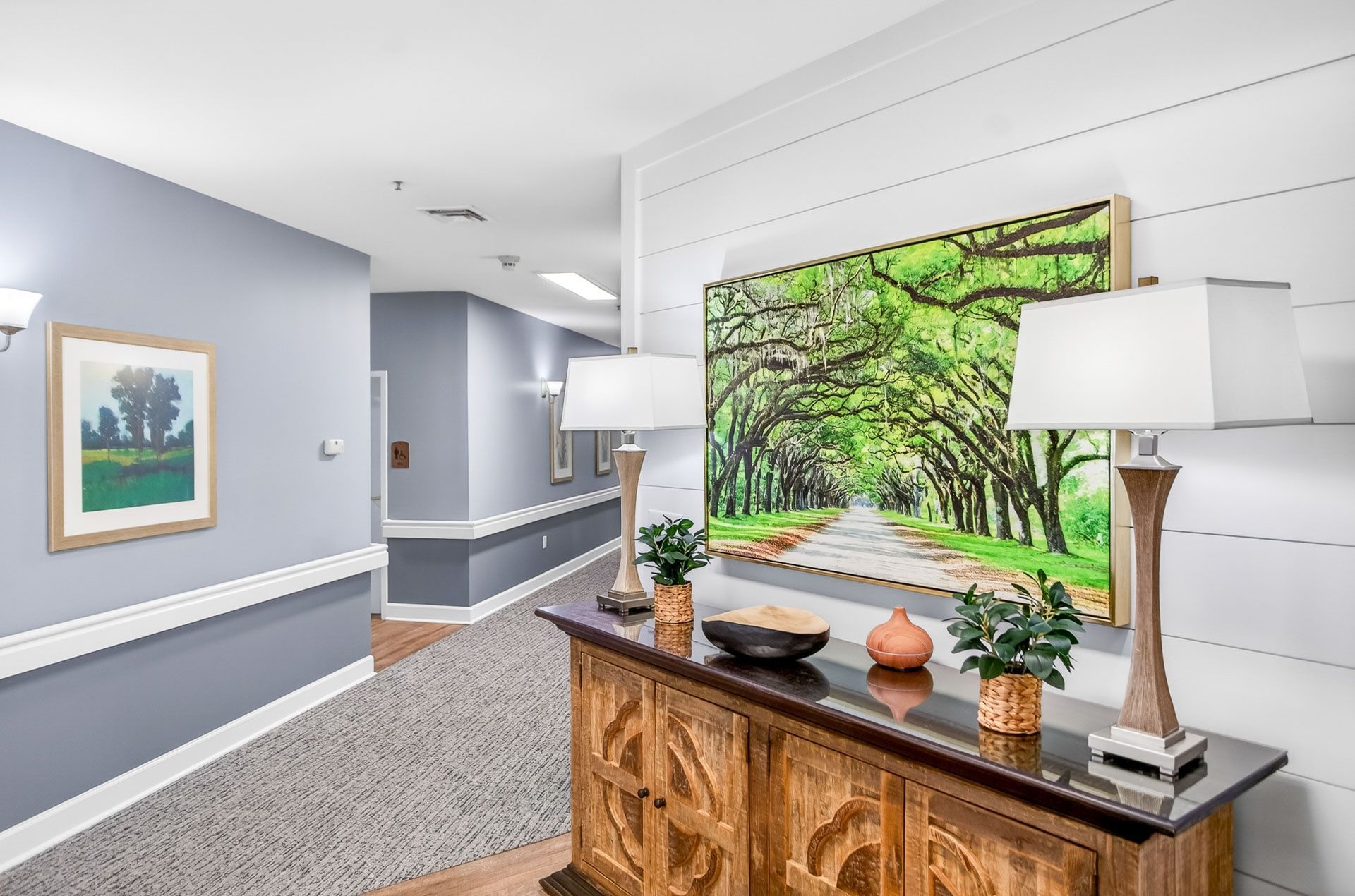 Bright hallway with white walls, framed art, a wooden console, lamps, and a large green landscape painting.