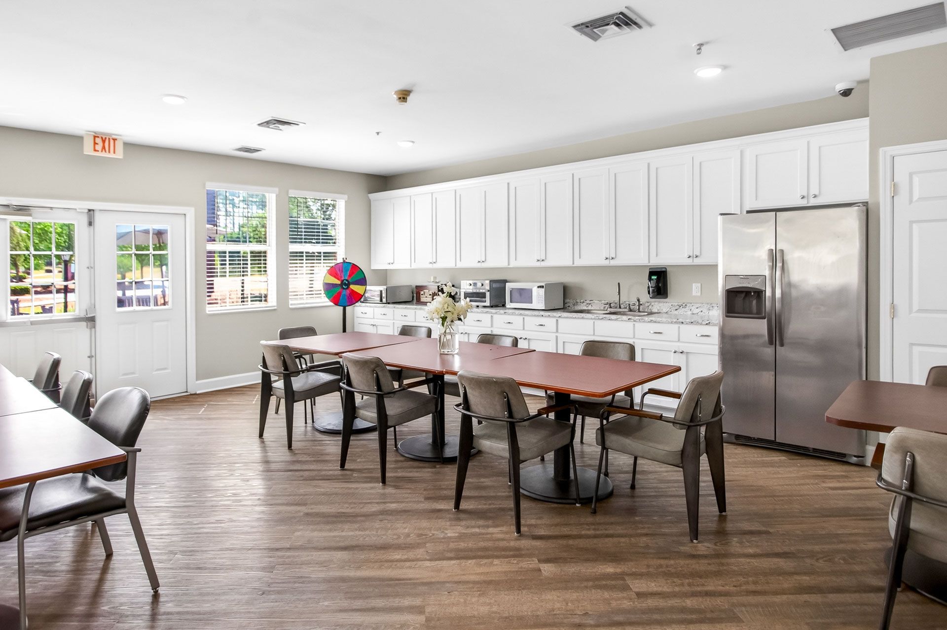 Bright community kitchen with long tables, white cabinets, stainless fridge, and glass doors to outside.