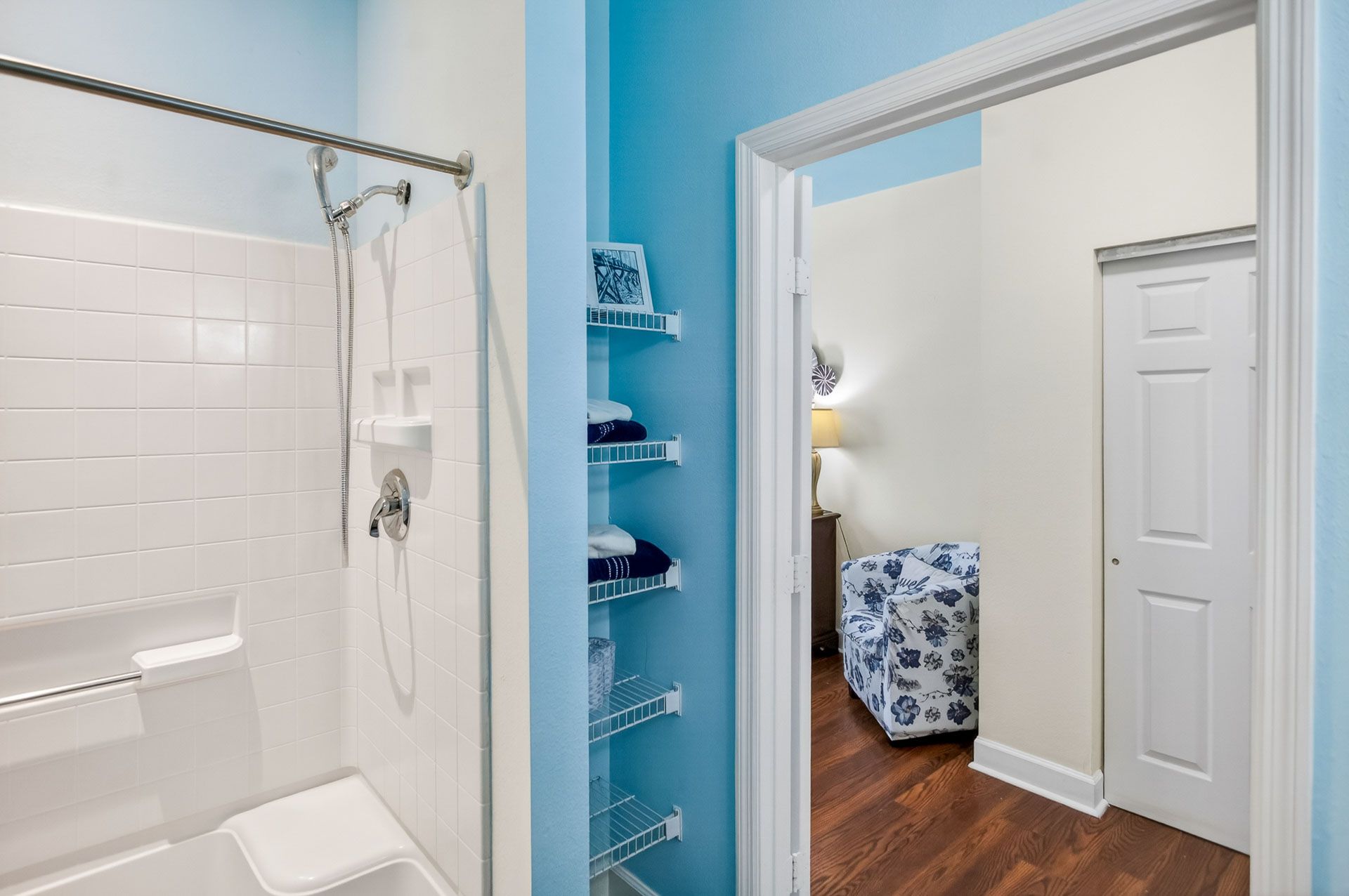 Bathroom with white tile shower, turquoise accent wall, and view into a bedroom with hardwood floors