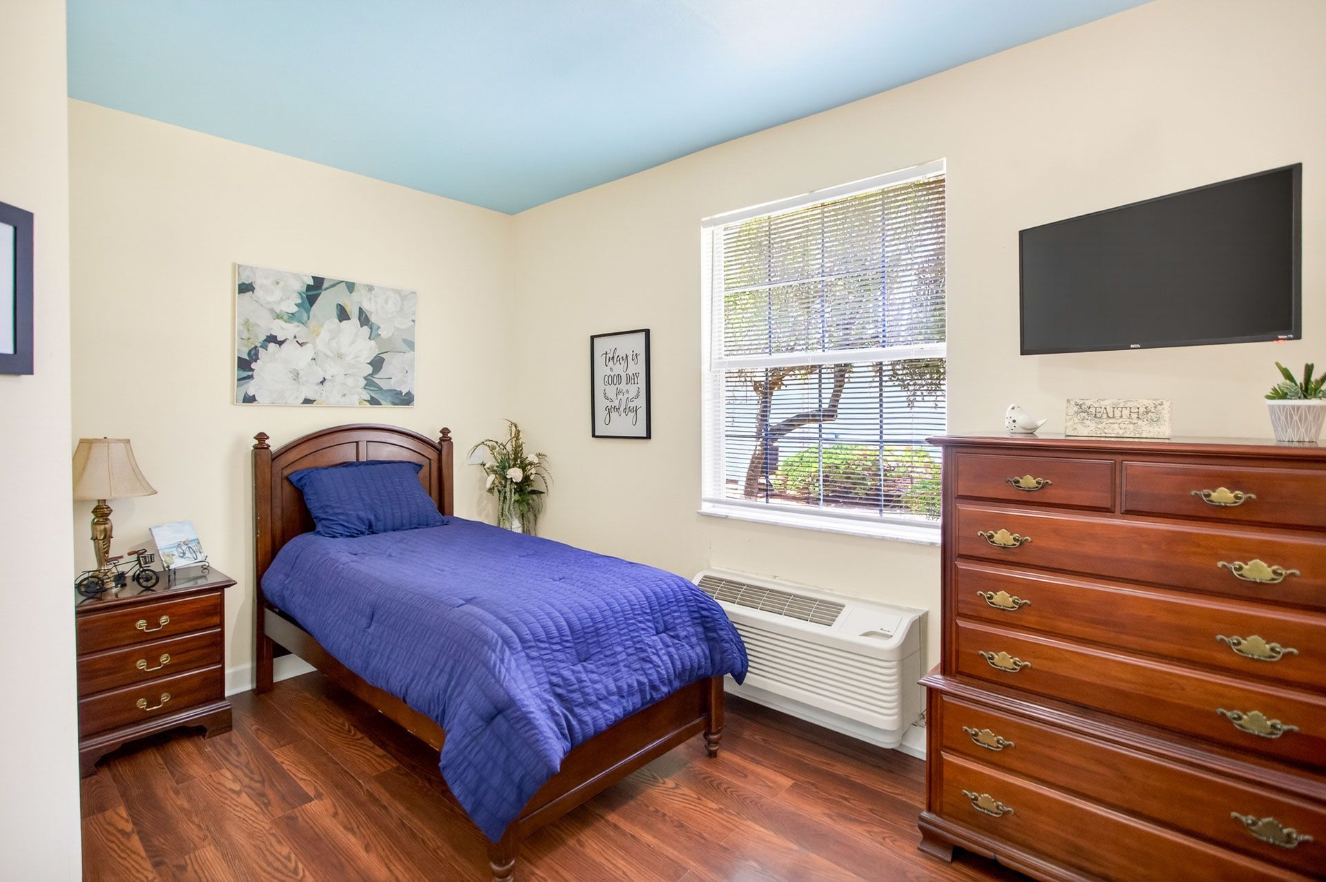 Small bedroom with blue bedspread, wooden dressers, wall TV, and a window with blinds