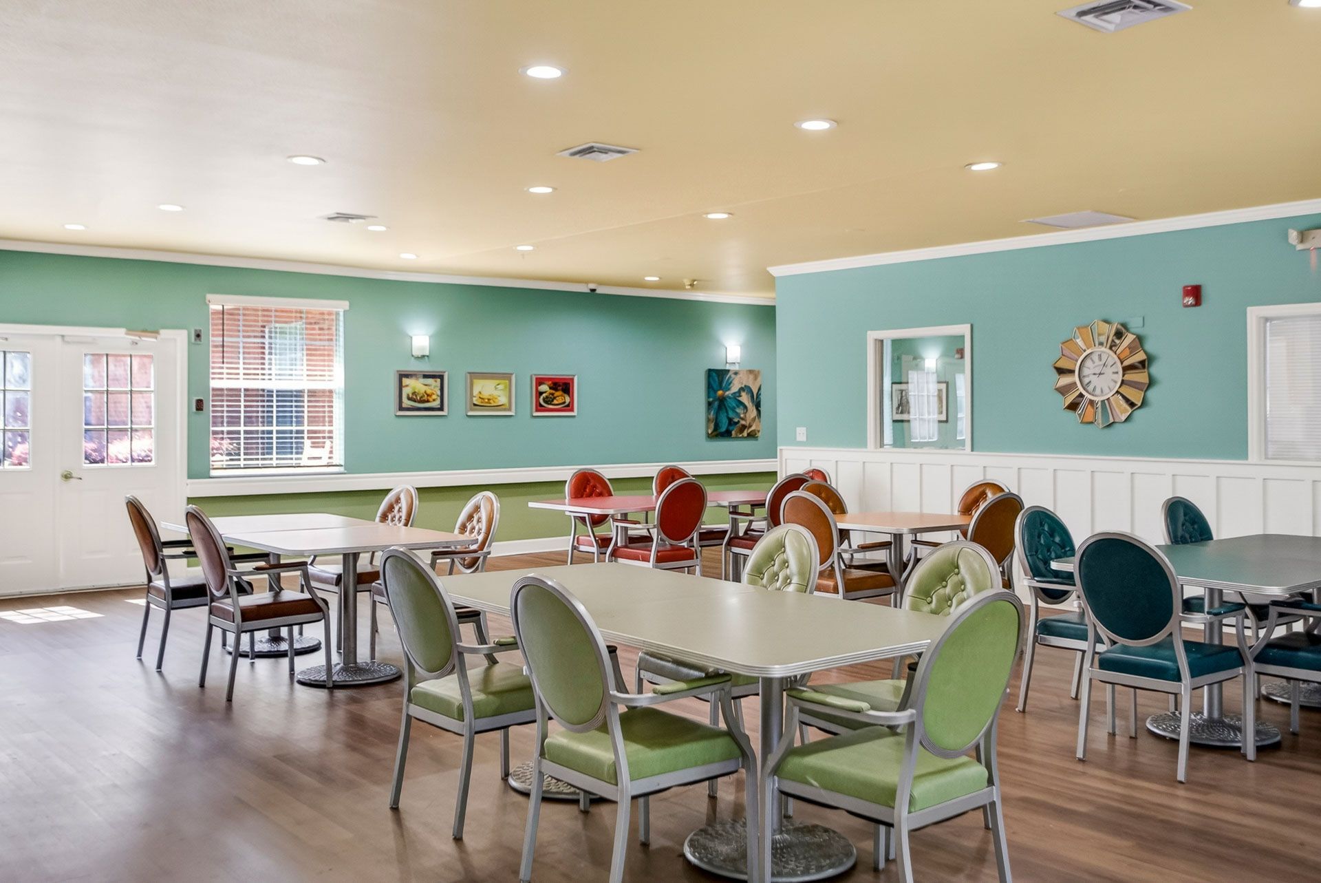 Bright dining room with colorful chairs, round tables, and teal walls in a senior living community