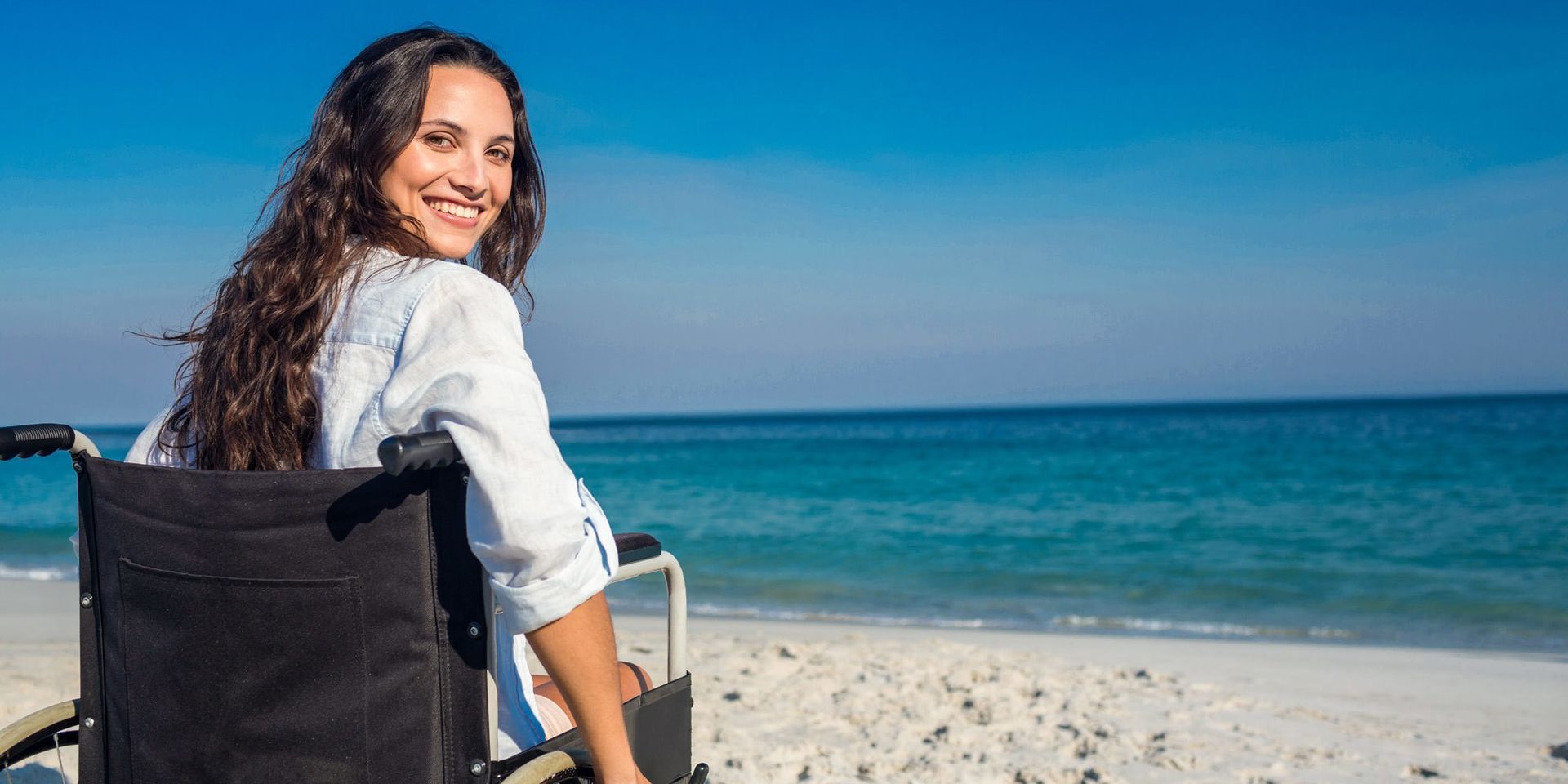 A woman in a wheelchair is sitting on the beach looking at the ocean.