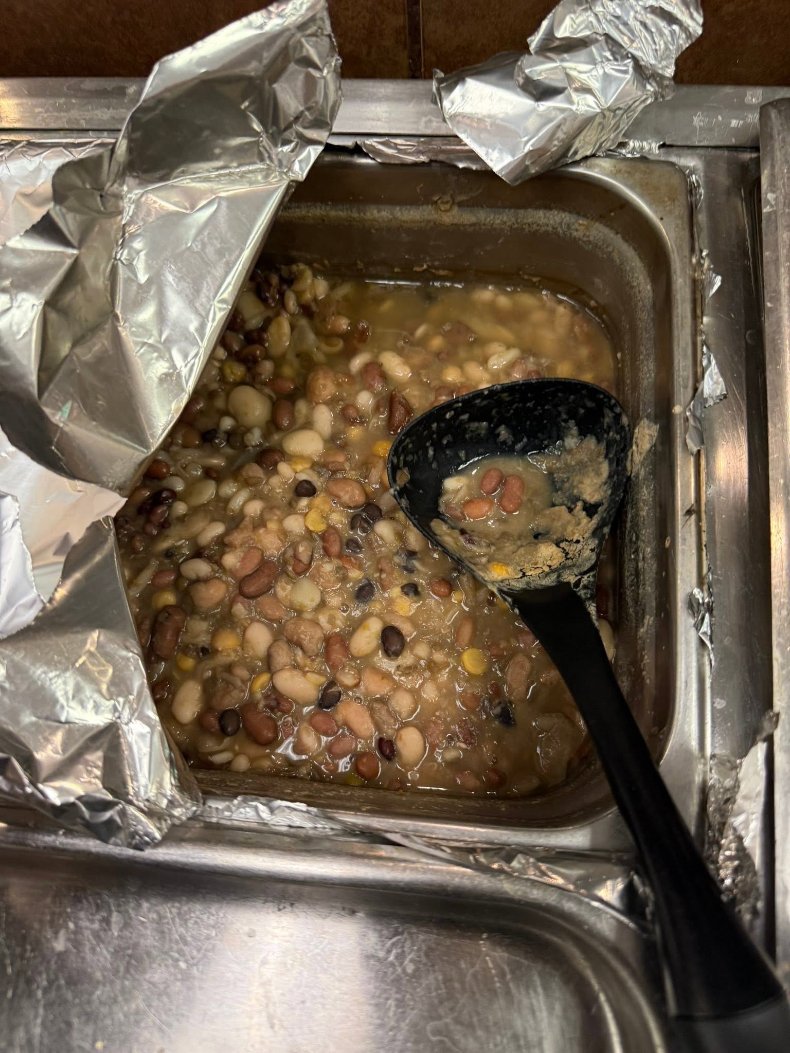 Foil-lined buffet tray with bean stew or soup and a black serving spoon inside
