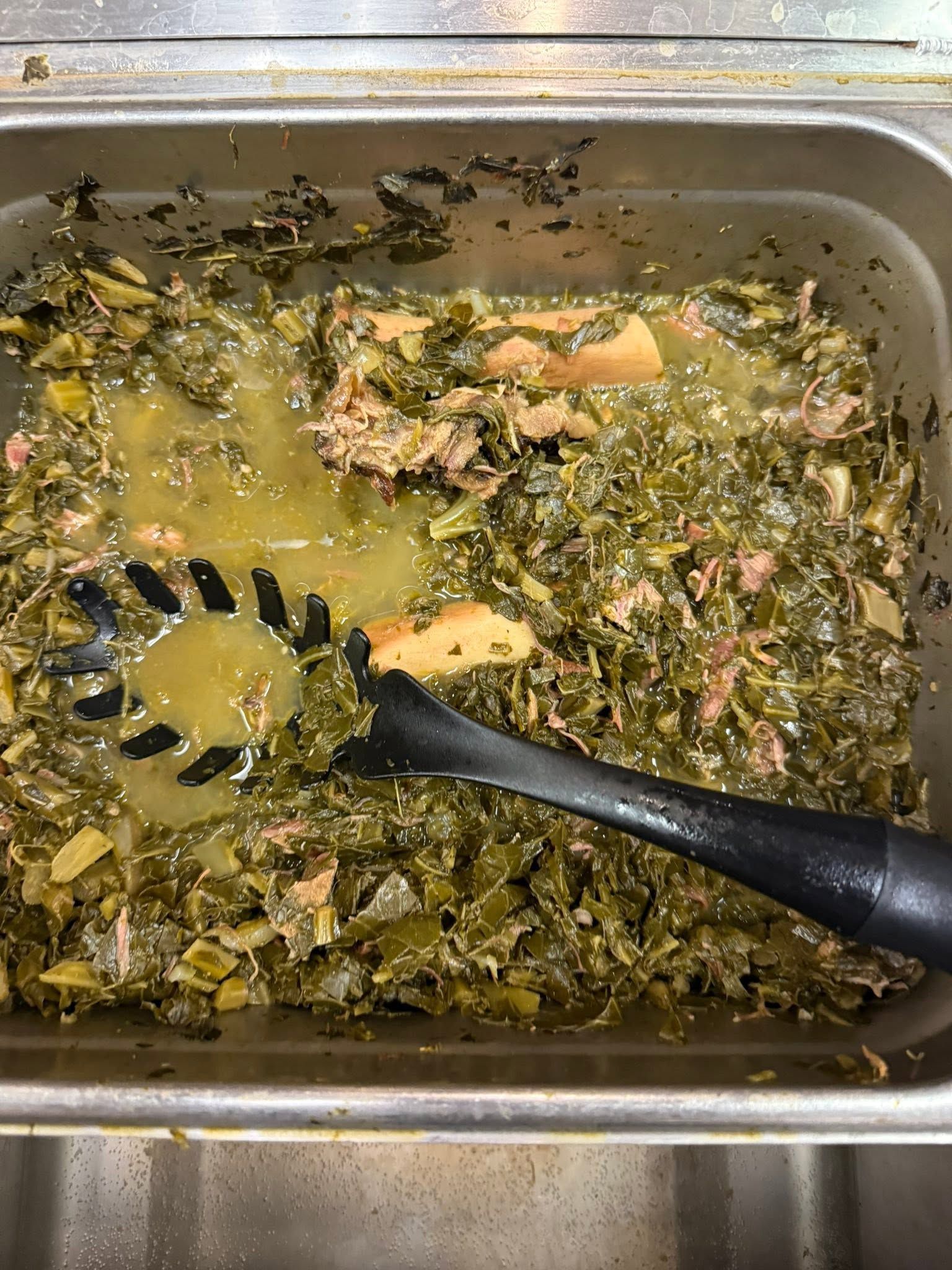 Messy tray of cooked greens in broth with a black serving spoon and a few orange pieces