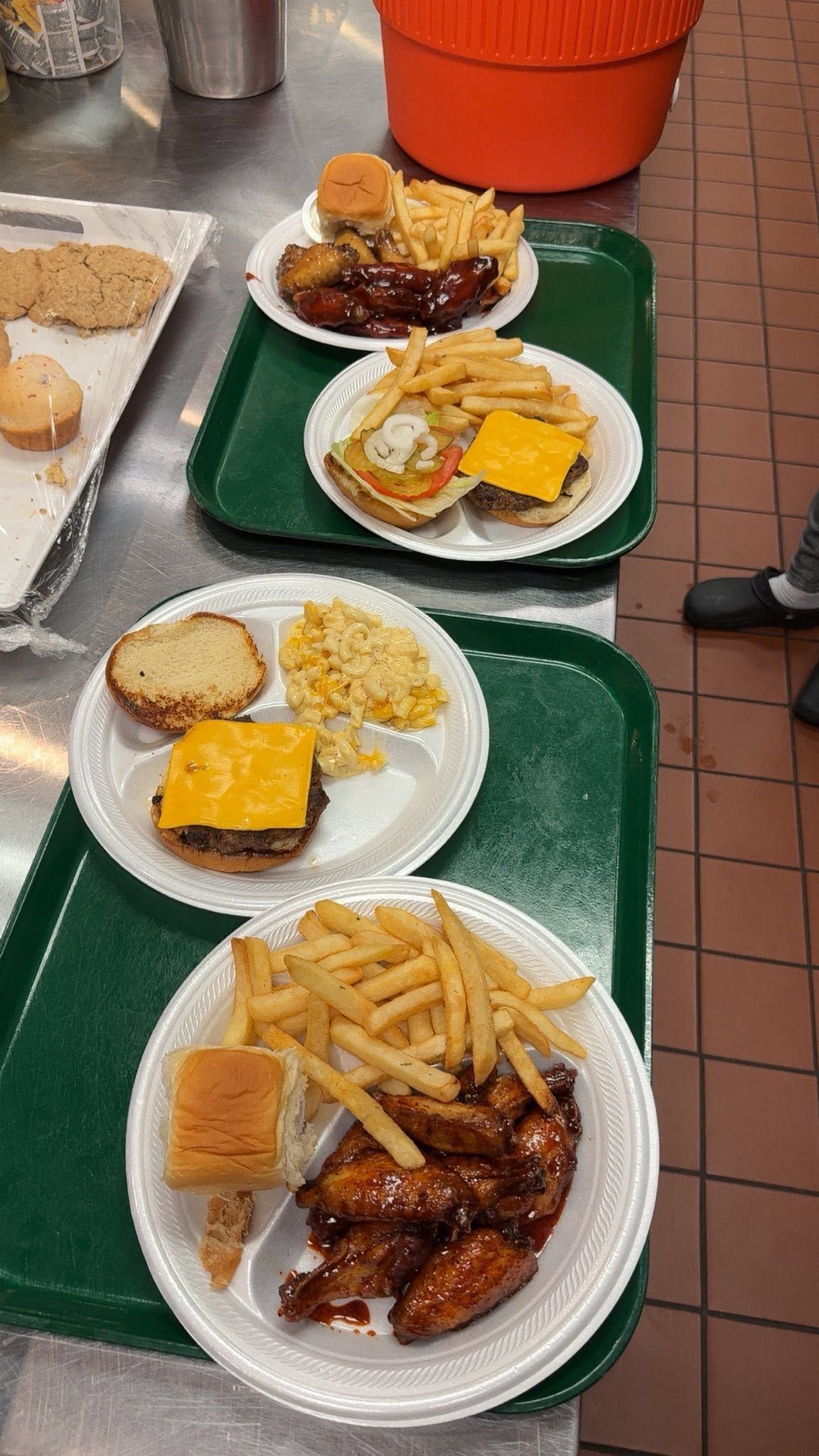 Three trays of cafeteria food with sandwiches, fries, and sauces on a tiled floor.