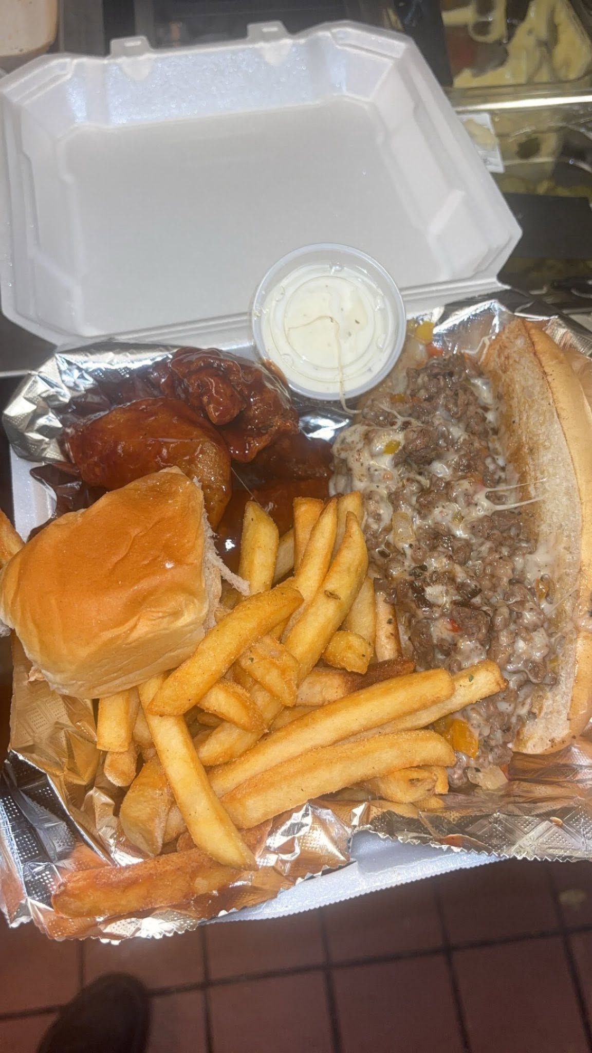 Takeout meal with sub sandwich, fries, roll, wings, and a small cup of dipping sauce in an open container