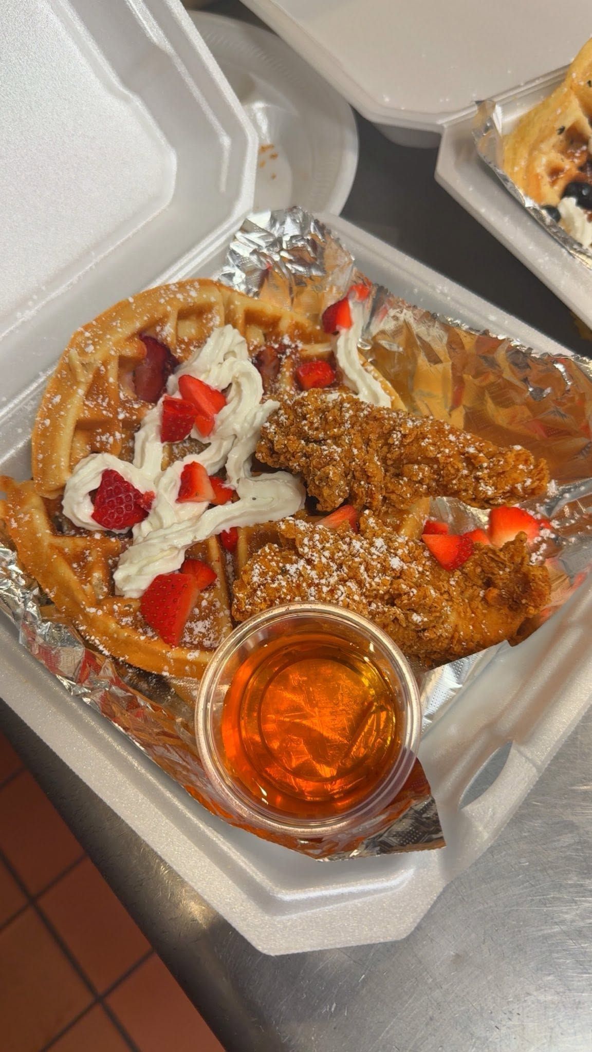 Foil takeout tray with syrup-soaked dessert topped with white cream and red pieces, in a foam container.