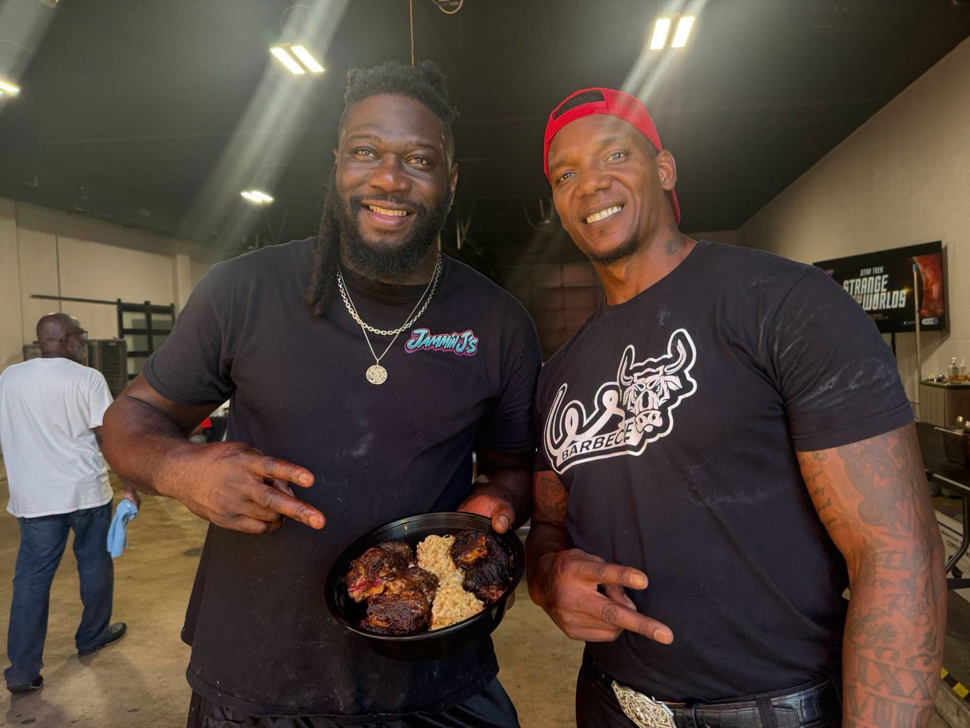 Two smiling men in black shirts pose indoors, holding a plate of grilled food.