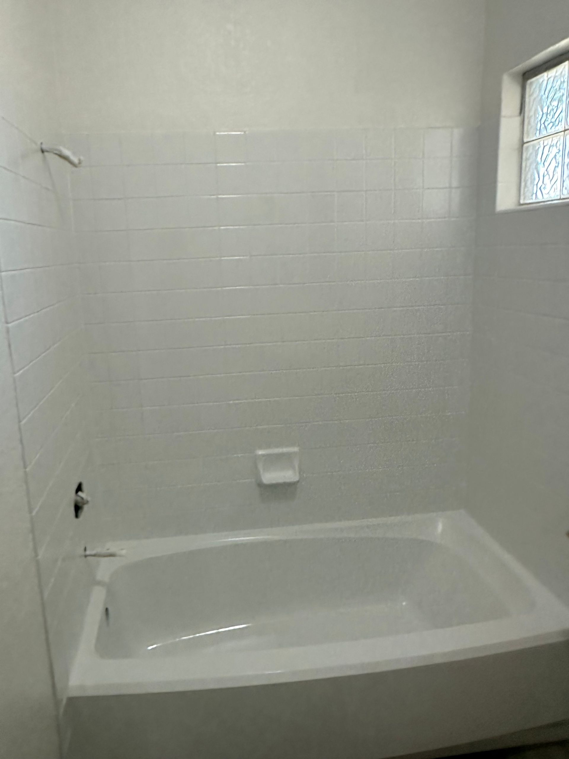 White-tiled bathroom with a bathtub, showerhead, and small window with stained-glass details.