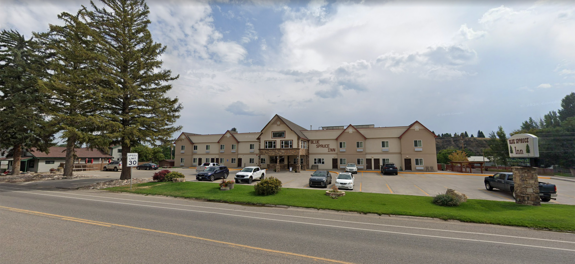 Street view of apartment buildings with parked cars, trees, and a cloudy sky