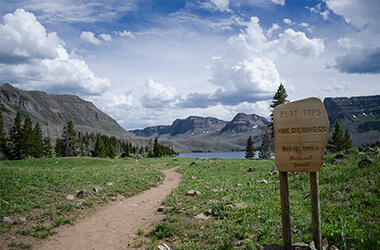 Mountain trail leading to a lake with rocky peaks under a cloudy sky; a sign stands at right.