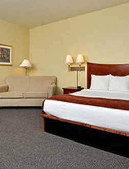 Hotel room with a red bed, white pillows, beige sofa, and two lamps on cream walls.