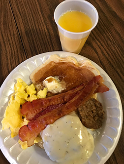 Breakfast plate with scrambled eggs, bacon, sausage, biscuit, gravy, and orange juice on a table
