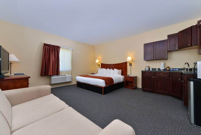 Hotel room with two beds, sofa, desk, and kitchenette in beige and dark wood tones
