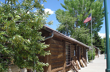 Wooden cabin beside a sidewalk, with trees and an American flag waving in the sun.