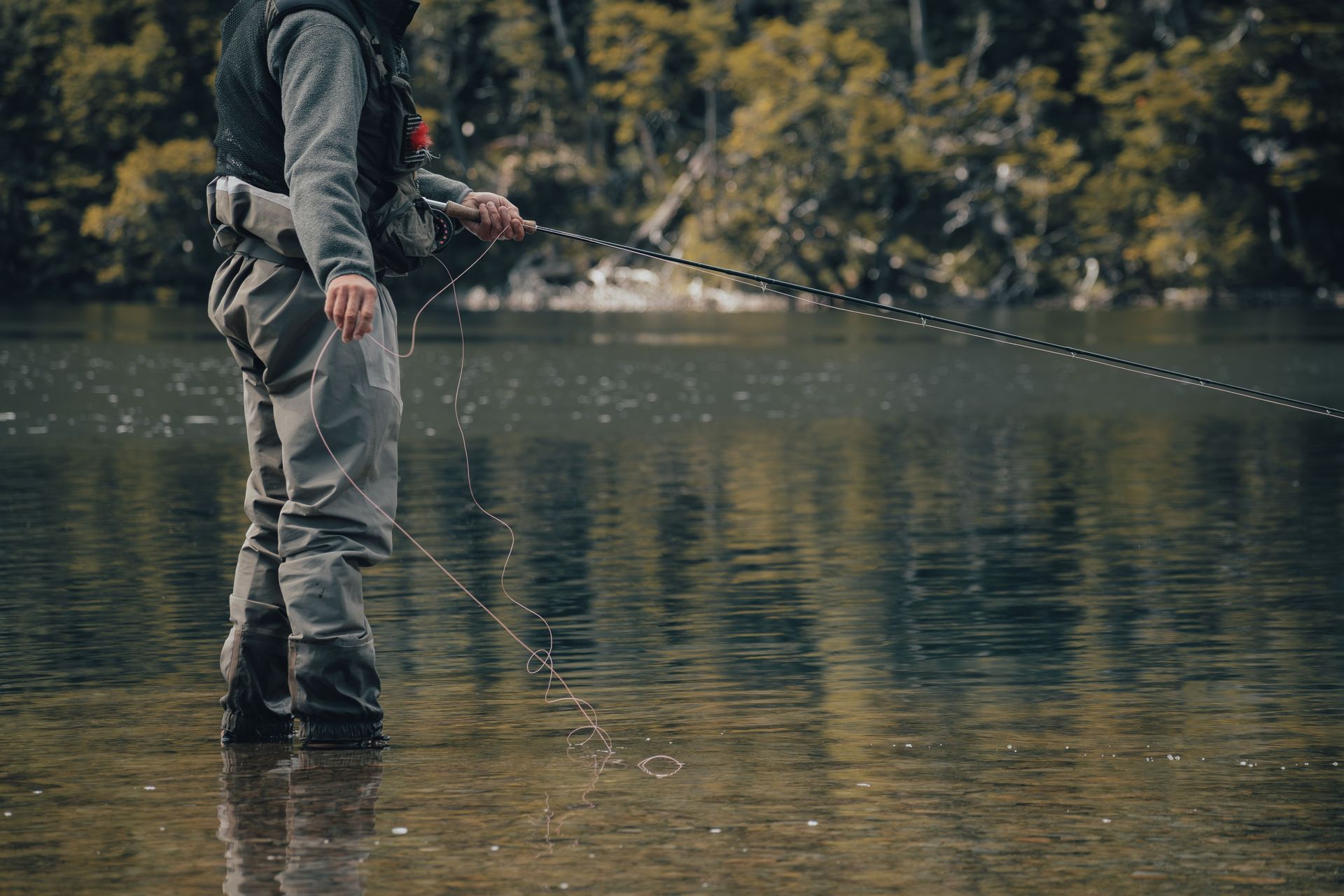 Person fly fishing in a calm lake, standing in shallow water with autumn trees in the background