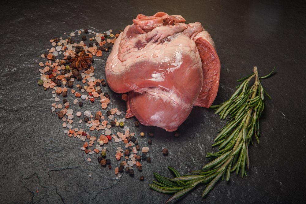 A raw pork heart surrounded by spices and rosemary on a table.