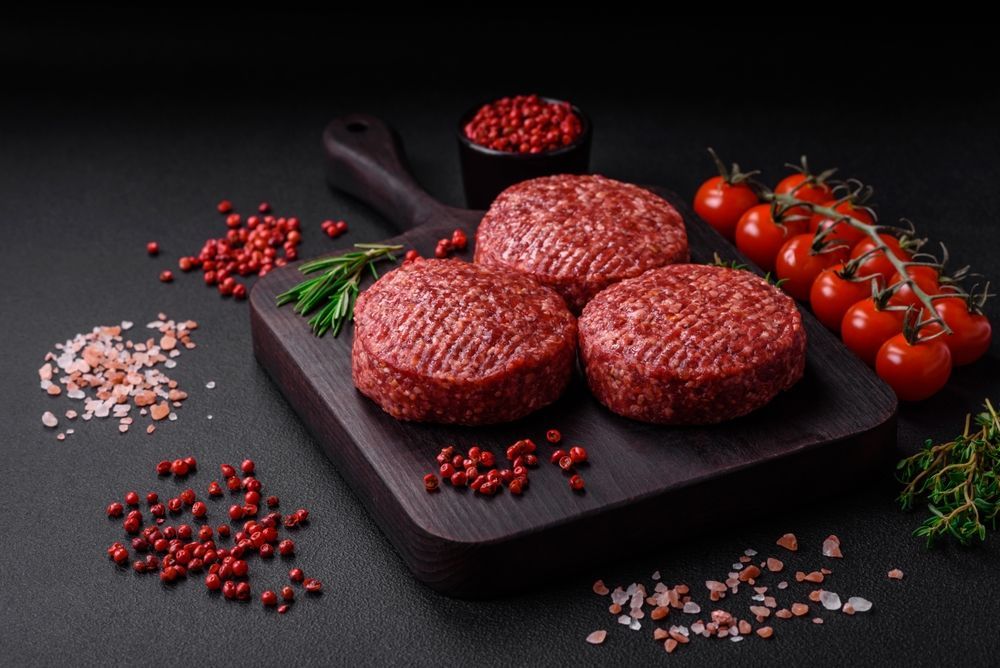 Three raw hamburger patties on a wooden cutting board with tomatoes and spices.