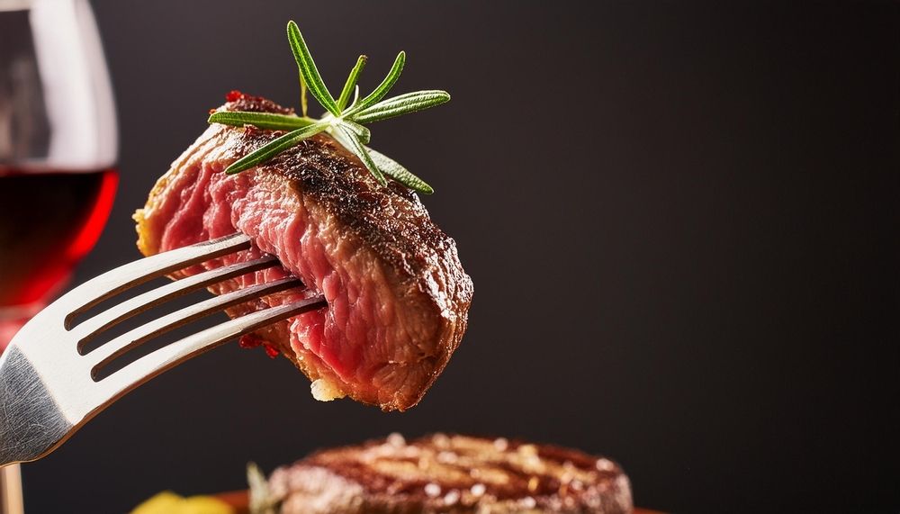 A piece of steak on a fork next to a glass of wine.