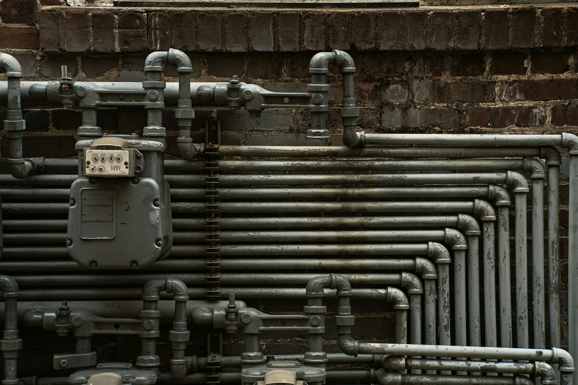 Industrial metal pipes and valves running along a brick wall in a utility setting