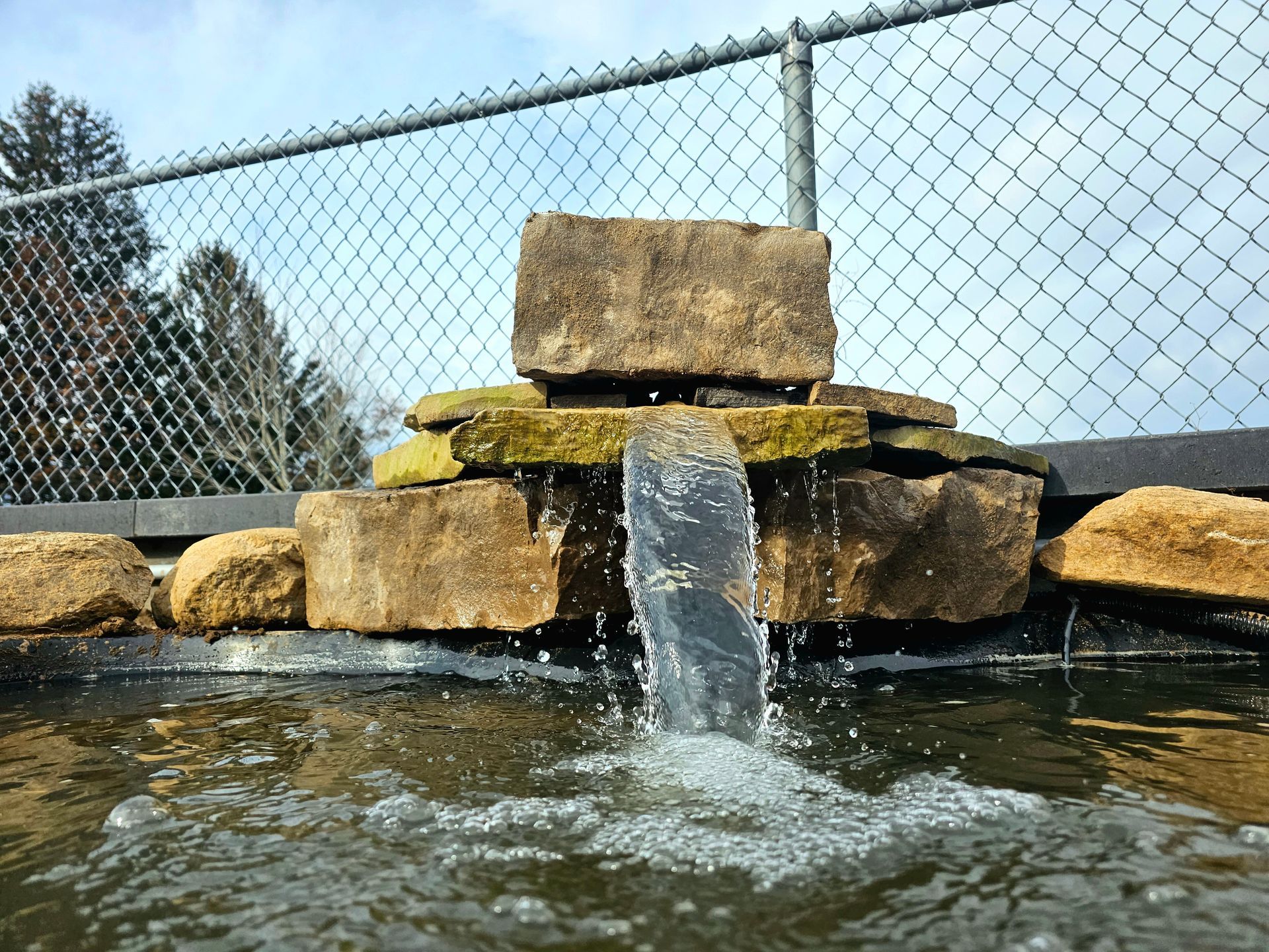 a waterfall in a pond with a chain link fence in the background .