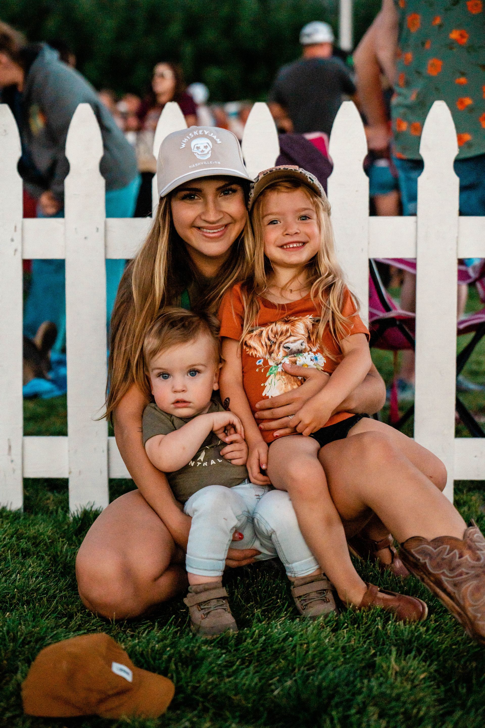 A woman is kneeling down with two children in front of a white picket fence.