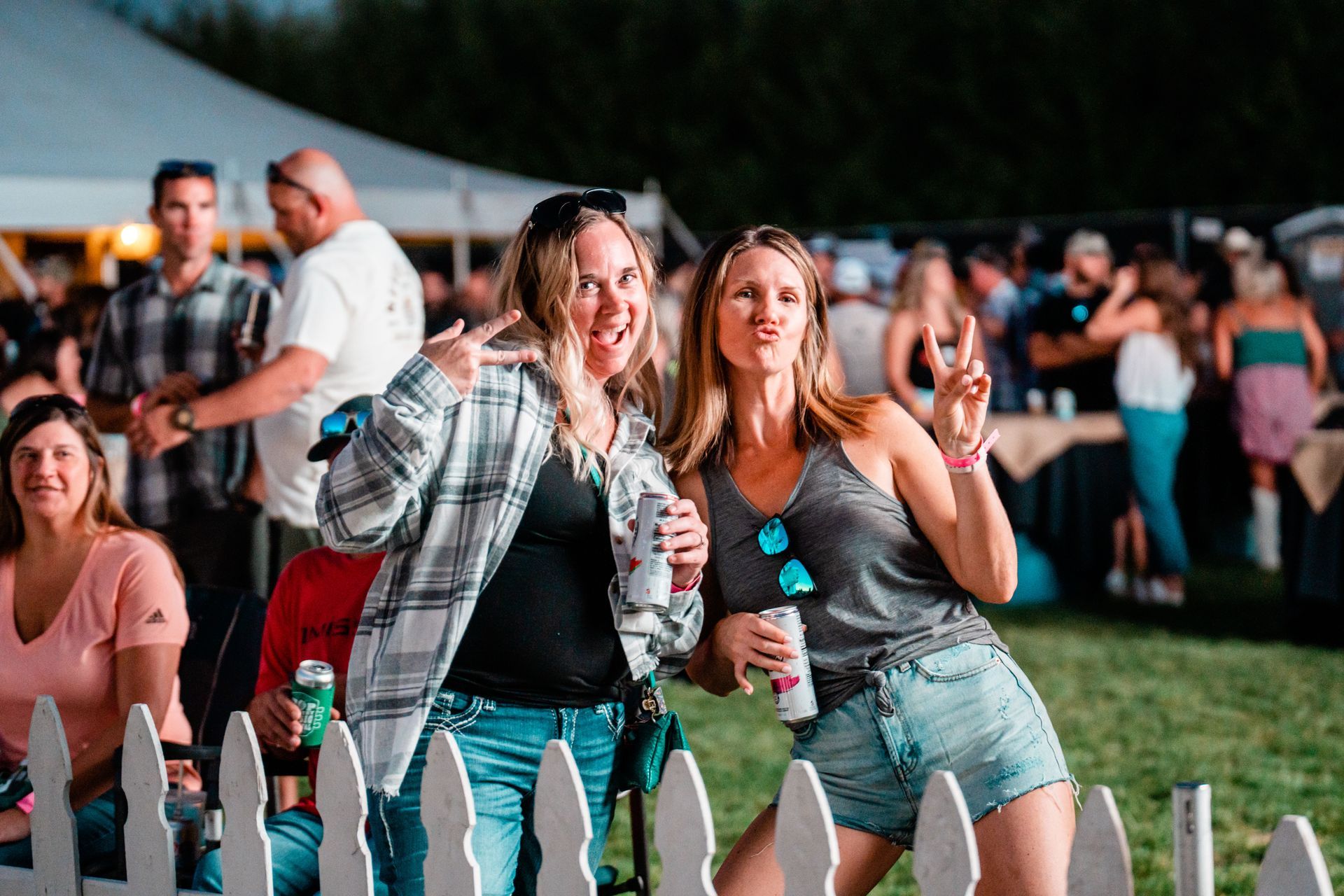 Two women are posing for a picture in front of a white picket fence at a festival.
