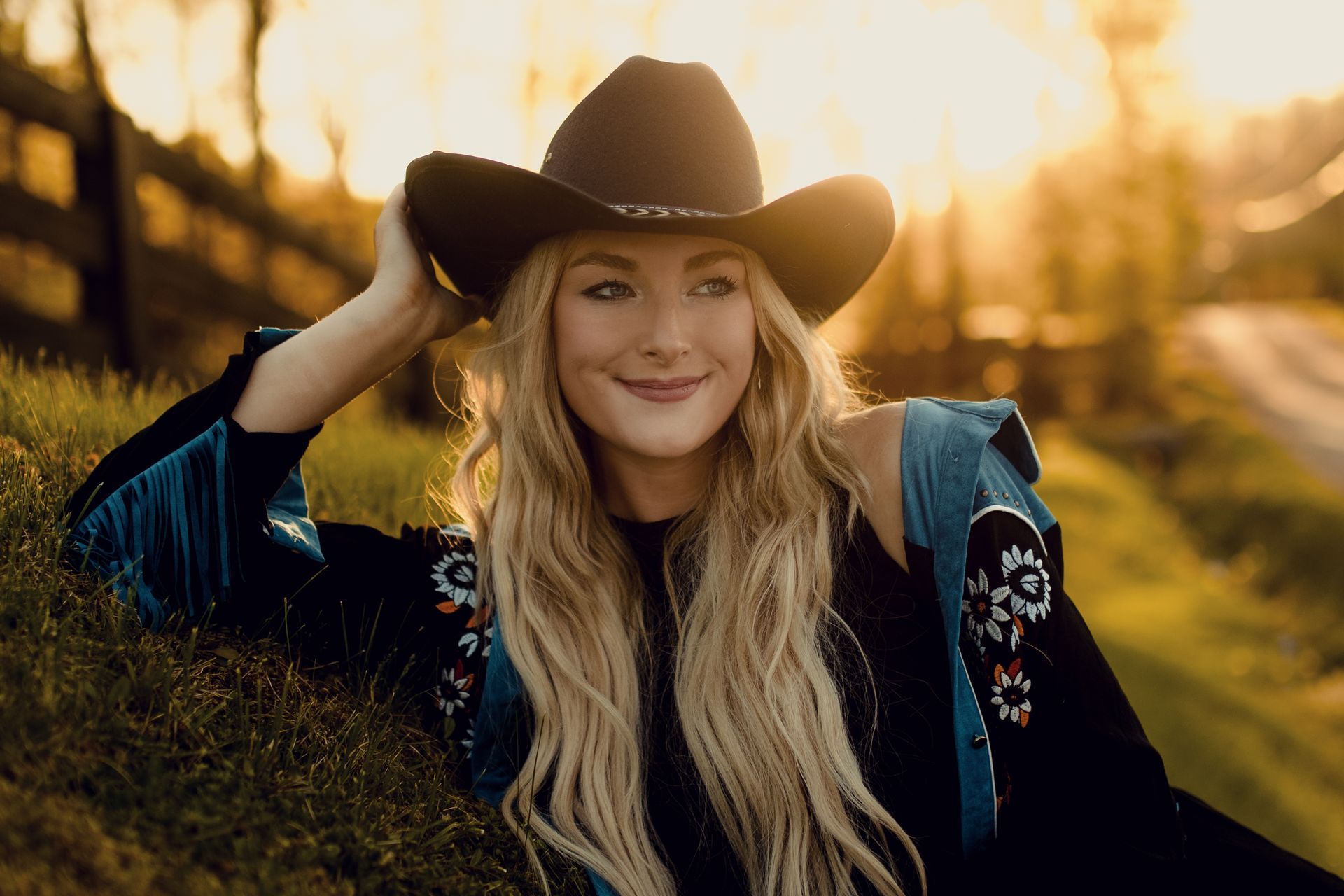 A woman wearing a cowboy hat and a blue jacket is laying in the grass.