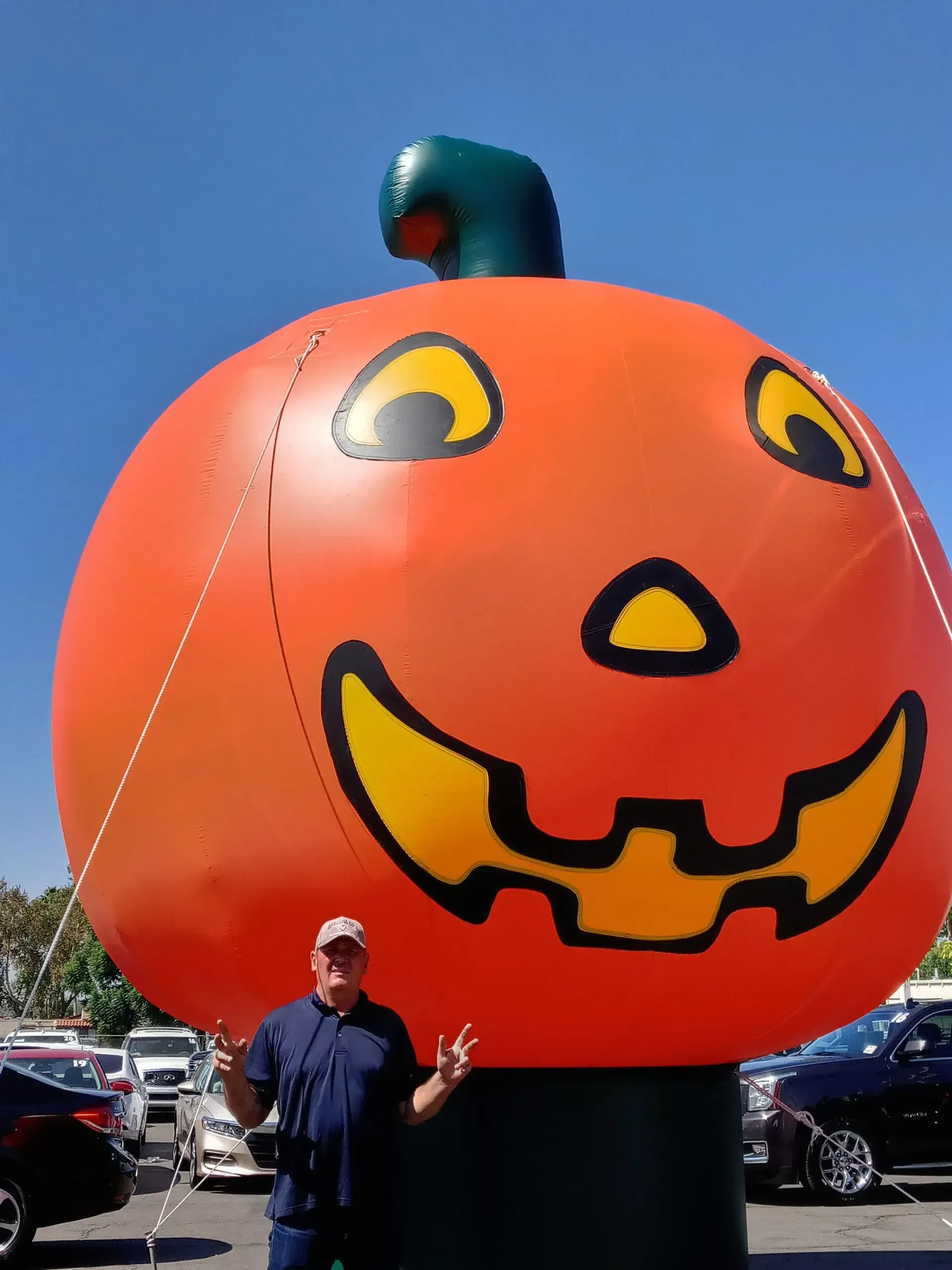 Large orange jack-o'-lantern decoration with a green stem in a parking lot, with a person standing below it