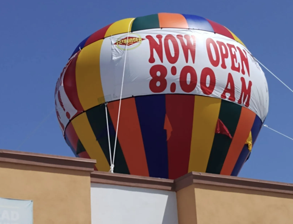 Large red-and-white “NOW OPEN 8:00 AM” hot air balloon sign above a building