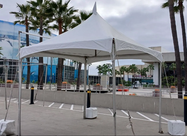 White event tent in an empty plaza beside palm trees and a glass building under cloudy skies