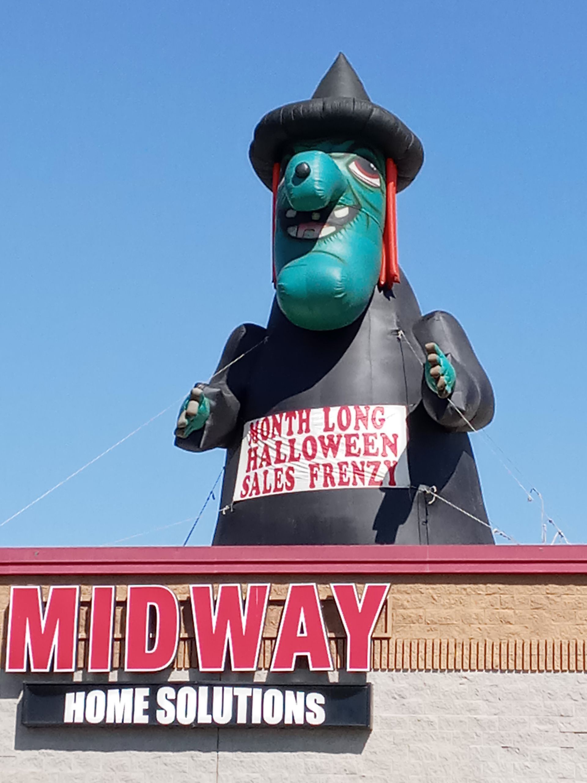 Large black-and-teal Halloween witch figure on a Midway Home Solutions sign under a red roofline.