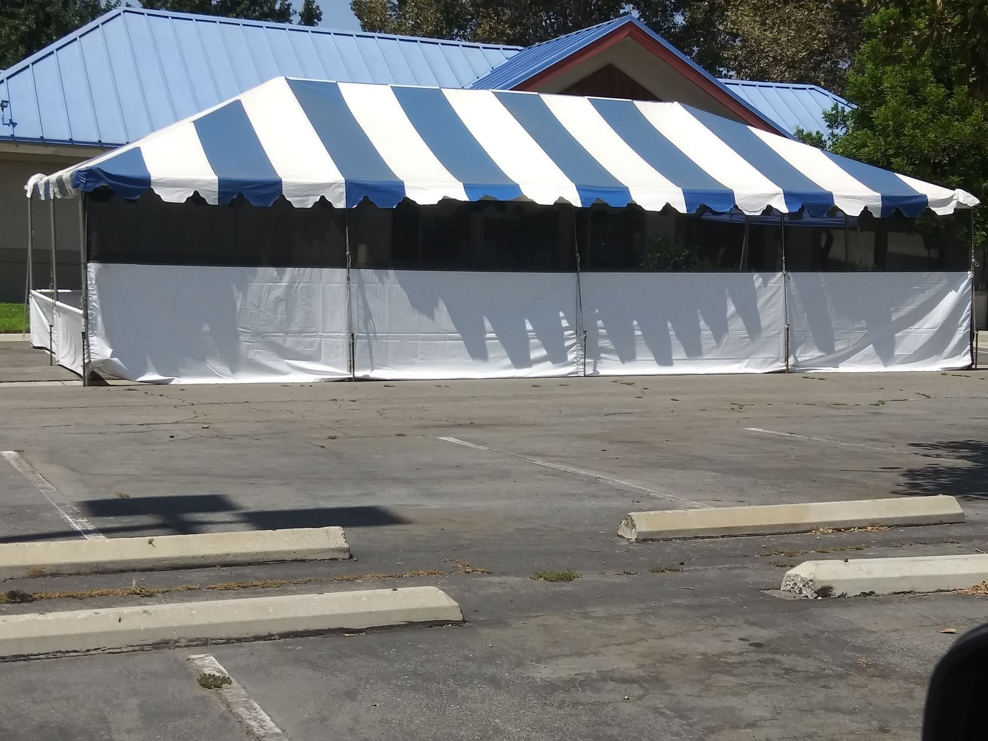 Blue-and-white striped canopy tent beside a parking lot, with white side panels and a building behind it