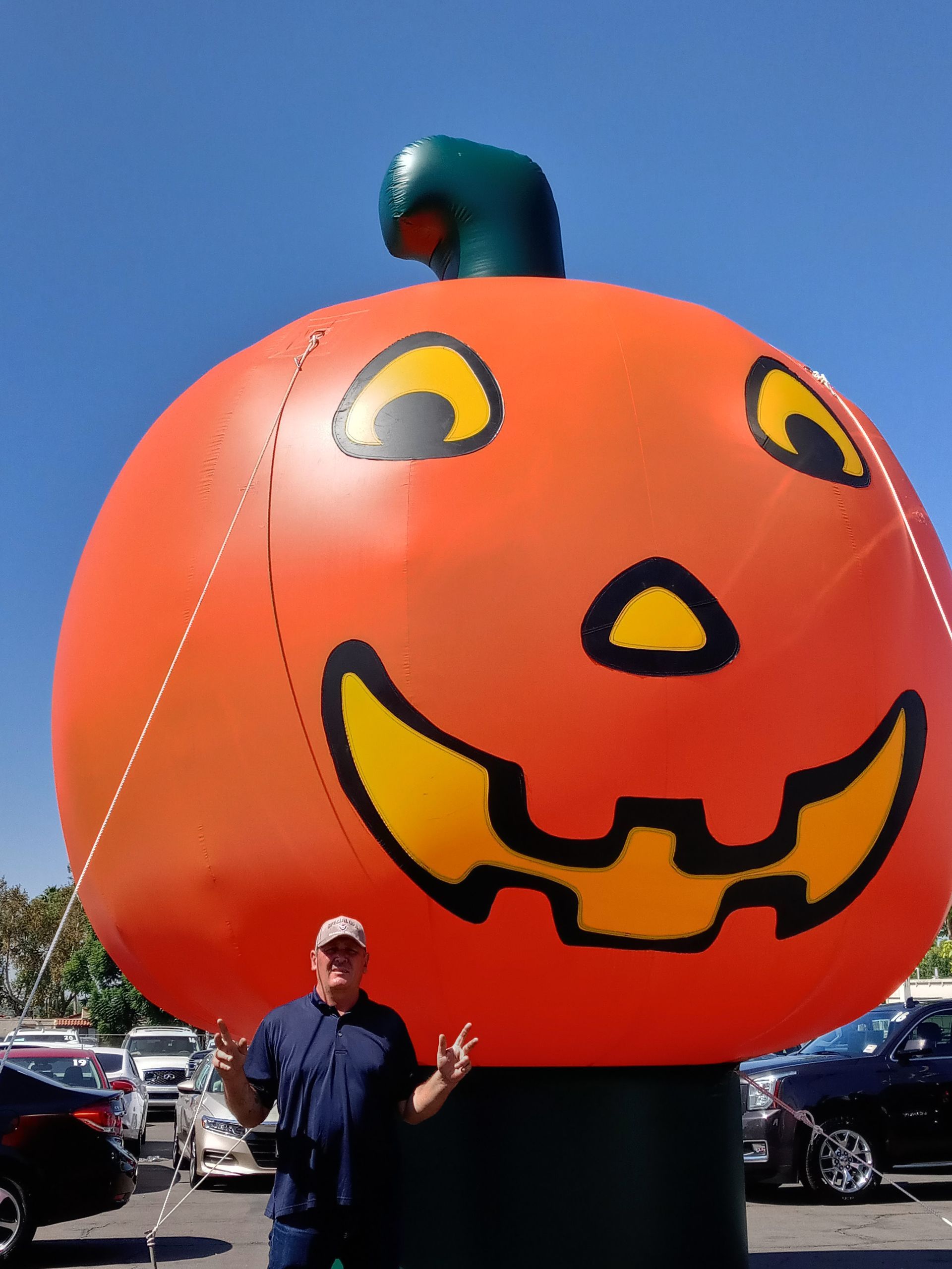 Large orange jack-o'-lantern display with a green stem, outdoors in a parking lot beside a person.
