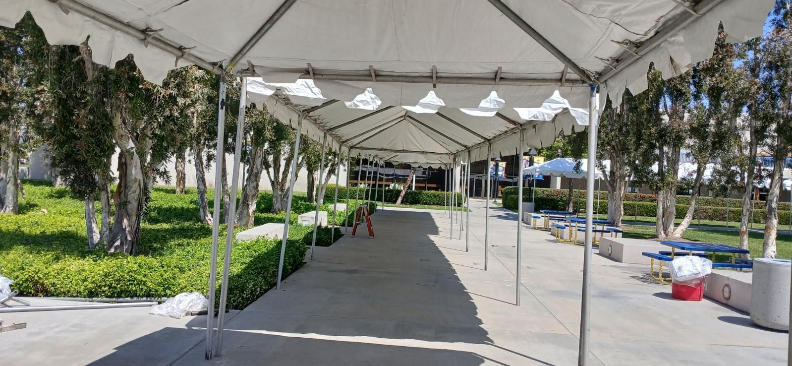 Covered outdoor walkway with white tents, trees, and people in the distance in a park-like setting