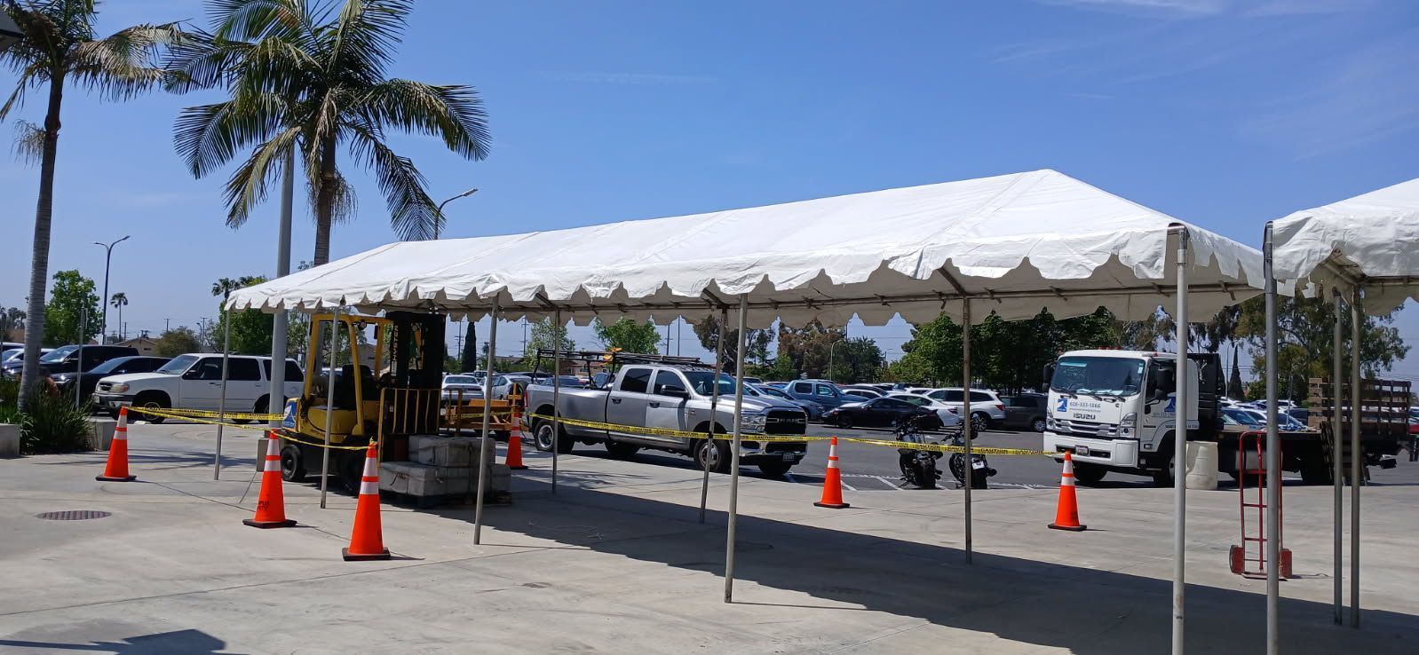 White event tent with orange cones in a parking lot near palm trees and parked cars