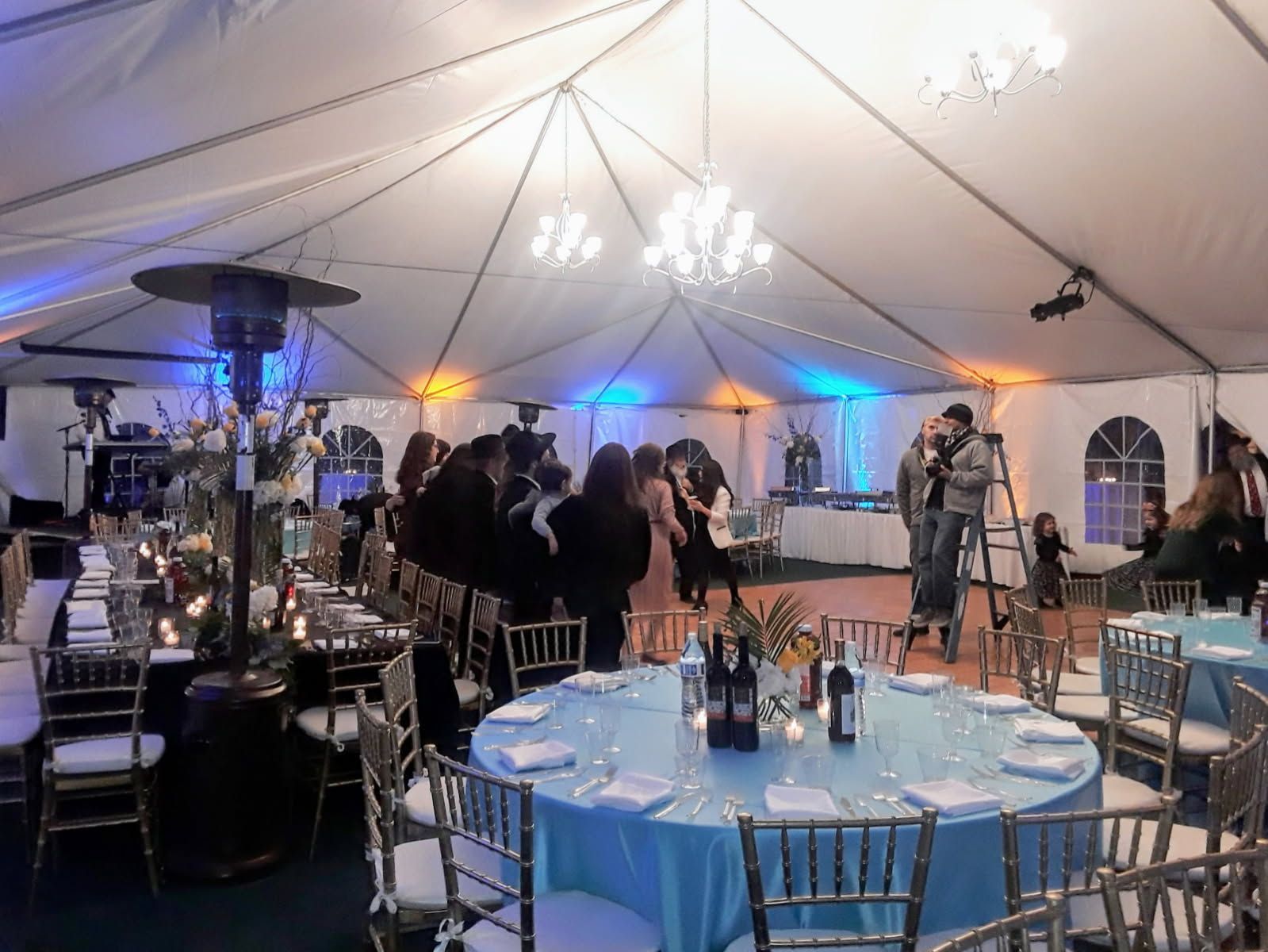 Guests mingle under a white event tent with chandeliers, blue lighting, and round tables set for a reception.