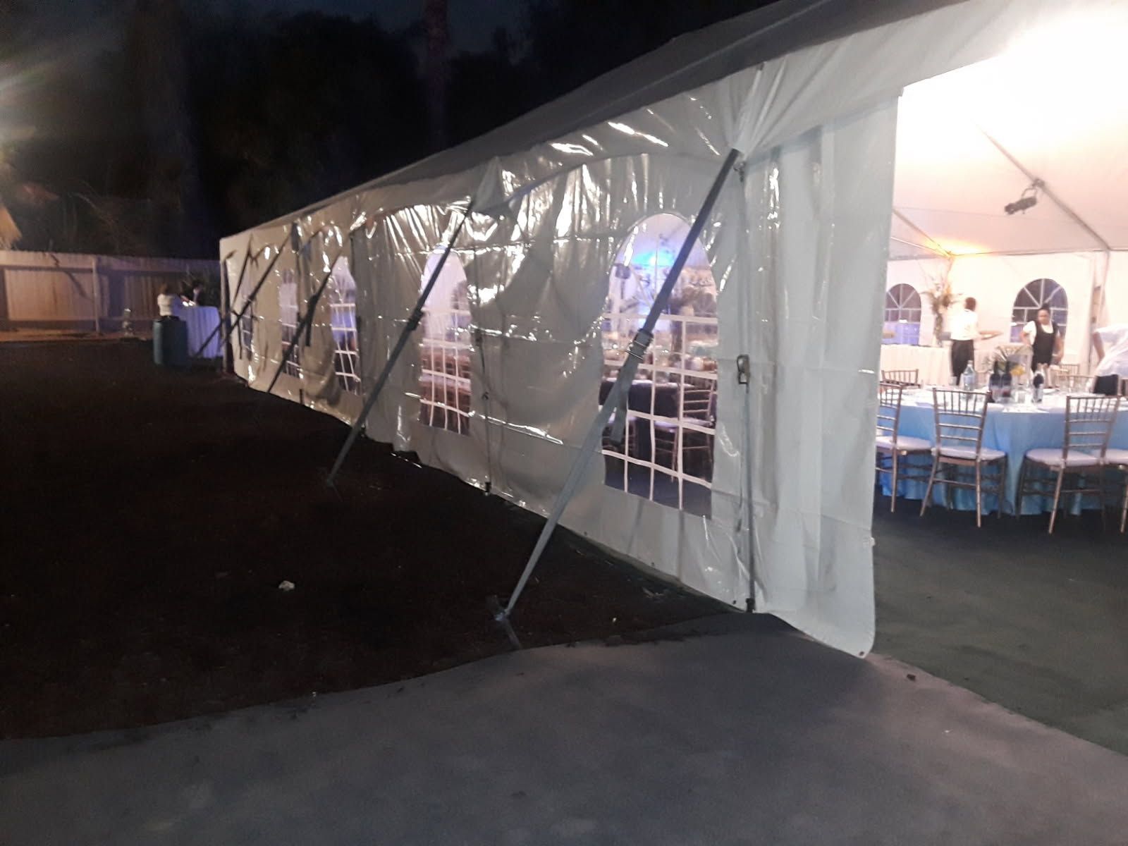 White event tent at night with tables and lights inside, viewed from outside along a paved area.