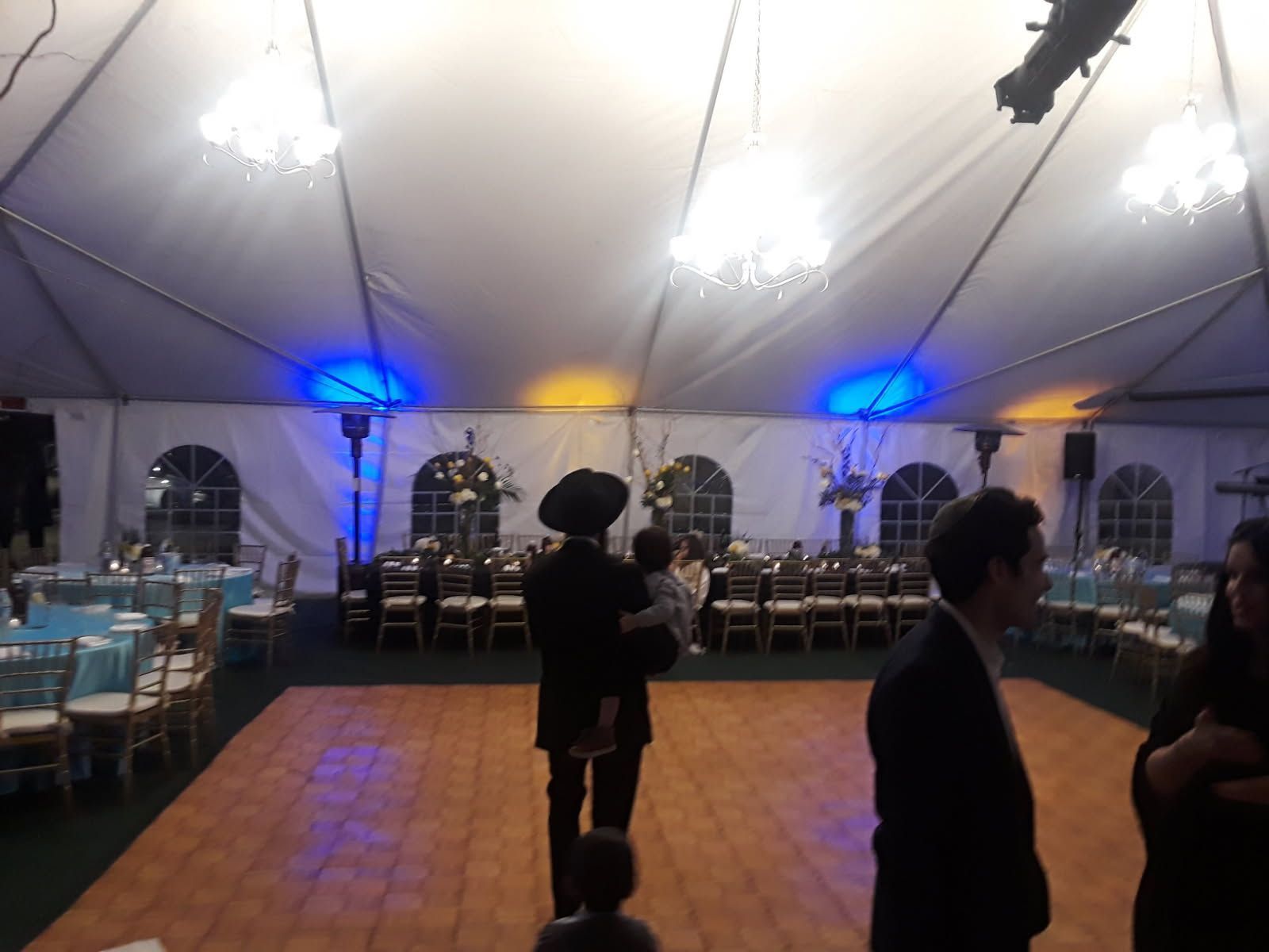 Indoor event tent with blue lights, dance floor, and banquet tables; a few people stand in the foreground.