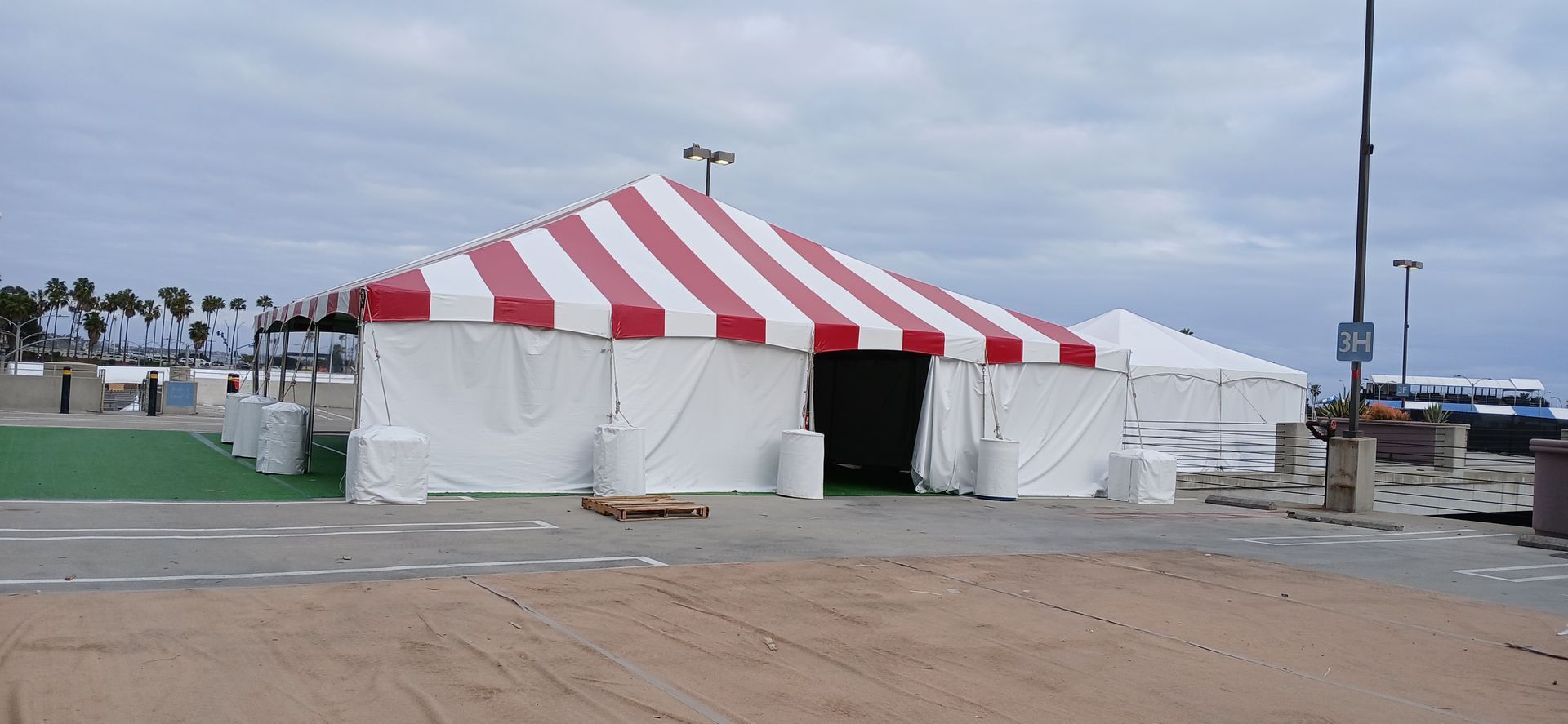 Red-and-white striped event tent on a sandy lot near the water under a cloudy sky