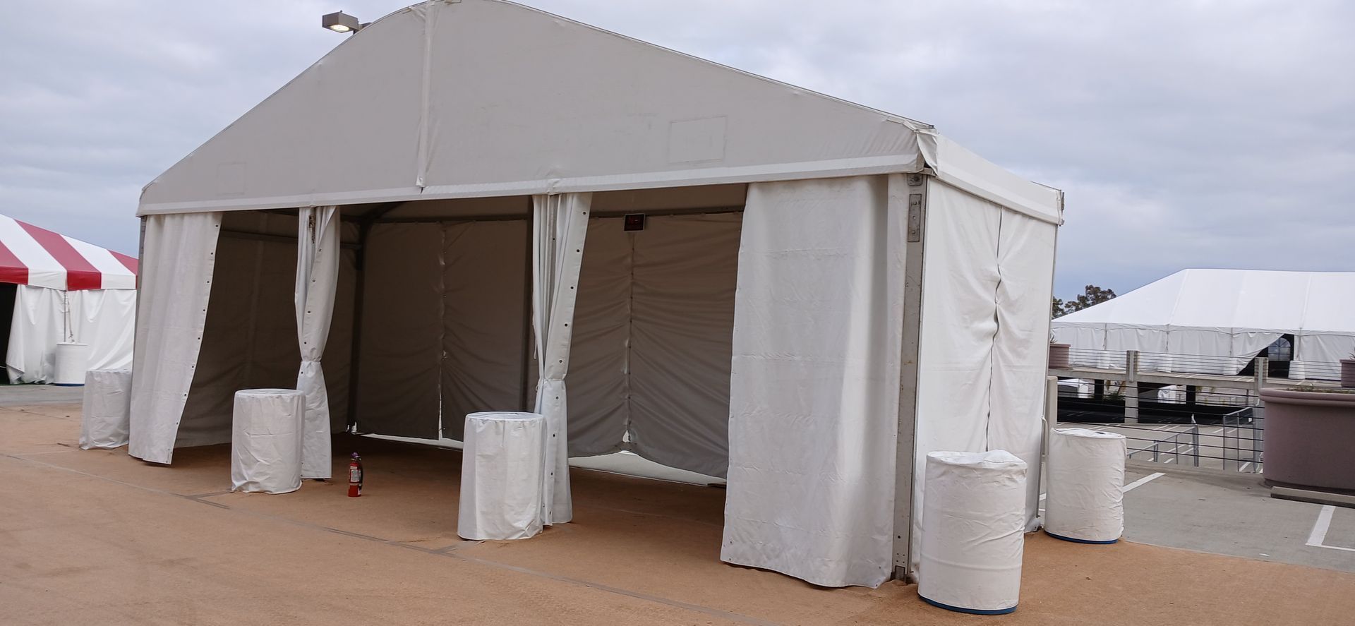 White event tent with open sides and beige curtains on a sandy outdoor lot