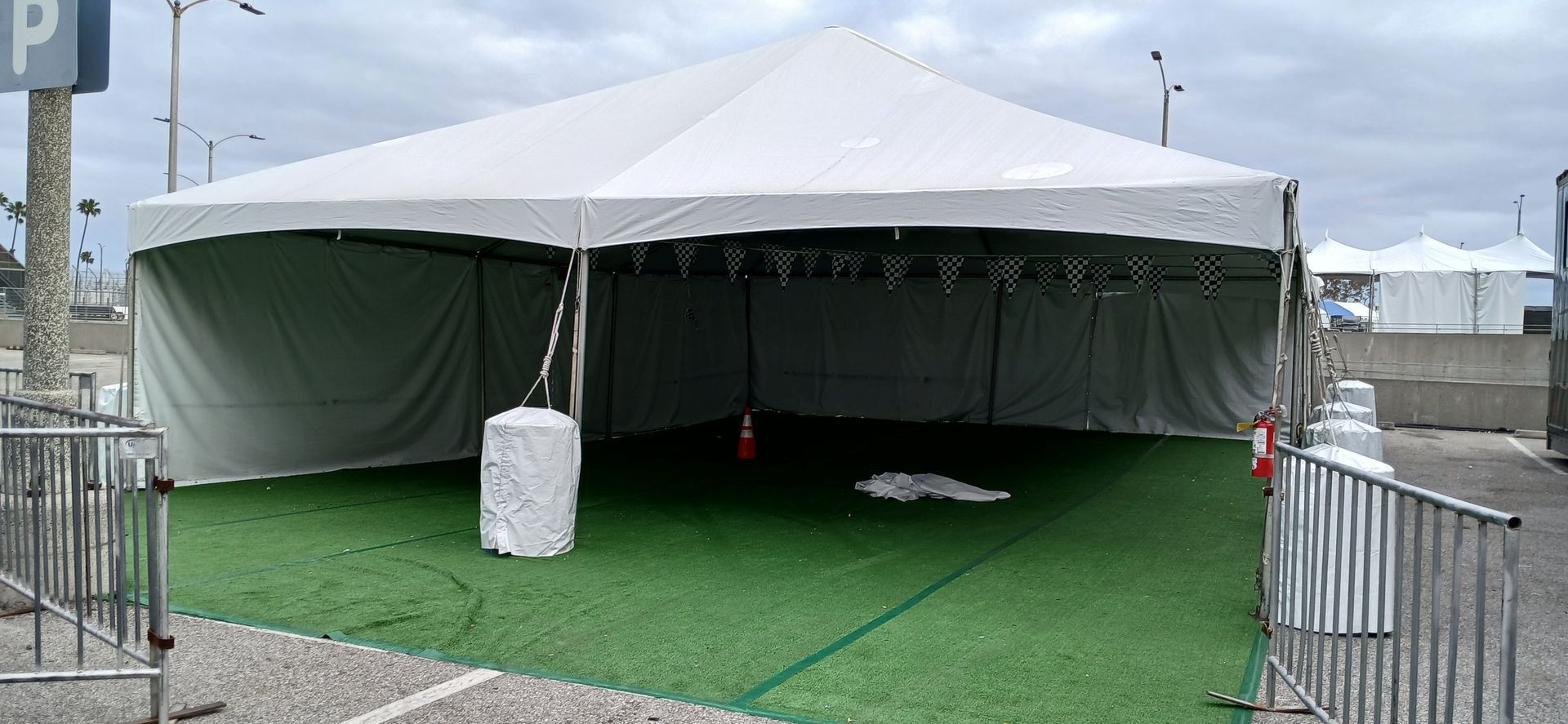 White event tent with open sides on green turf, surrounded by metal barricades under an overcast sky