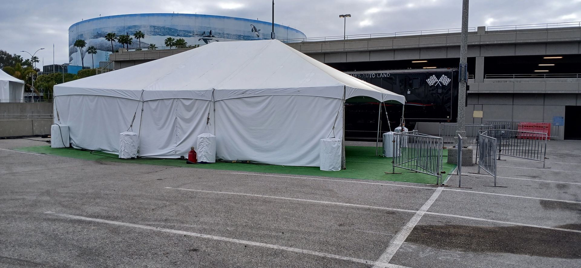 White event tent beside a parking lot and overpass, with green fencing and a red cone nearby