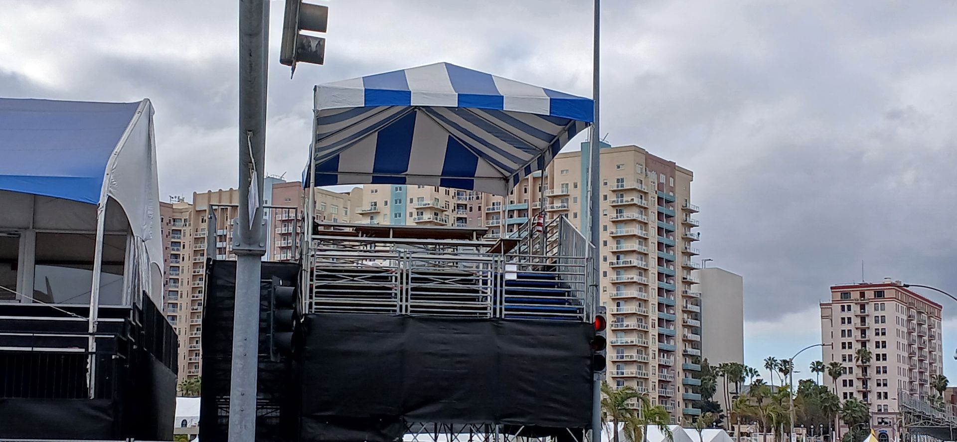 Blue-and-white striped canopy over a stage setup with city buildings in the background under cloudy sky