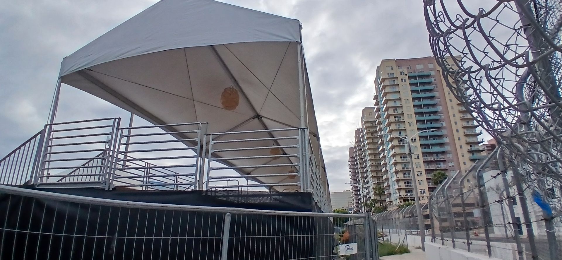 Covered outdoor pavilion beside a fence, with tall apartment buildings in the background on a cloudy day