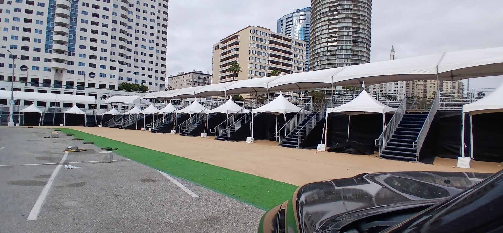 Row of black event tents beside a green carpet in a city parking lot with tall buildings nearby