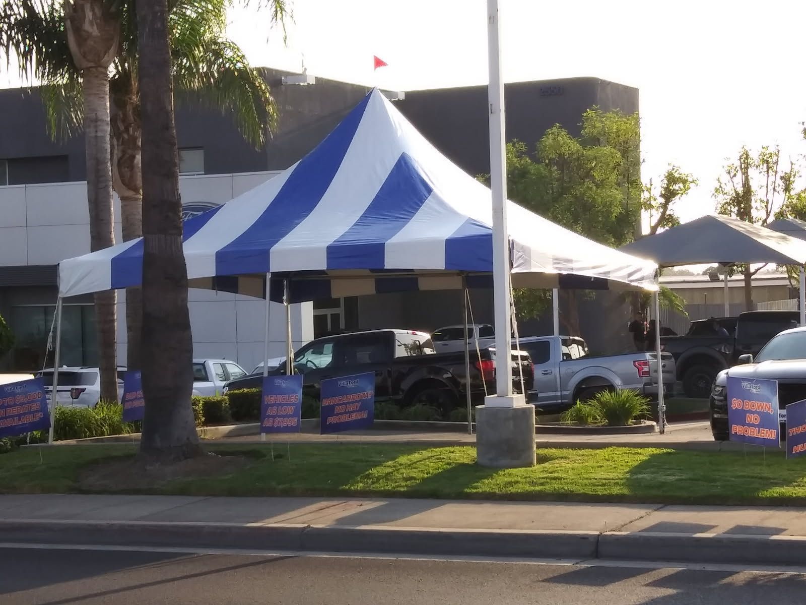 Blue-and-white striped event tent beside palm trees and parked cars on a sunny street corner