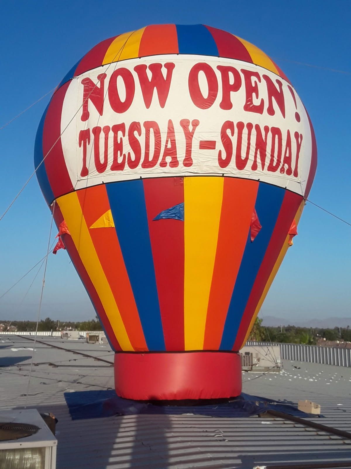 Colorful hot air balloon sign reading “NOW OPEN TUESDAY-SUNDAY” on a rooftop under a clear sky